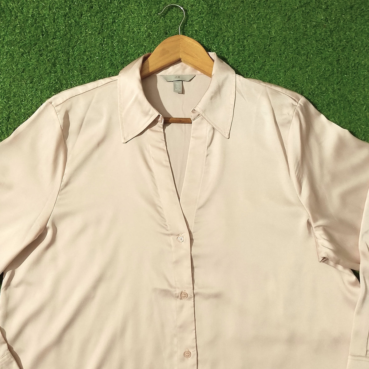 A light beige long-sleeve button-up shirt with a pointed collar, hanging on a wooden hanger against a green artificial grass background.