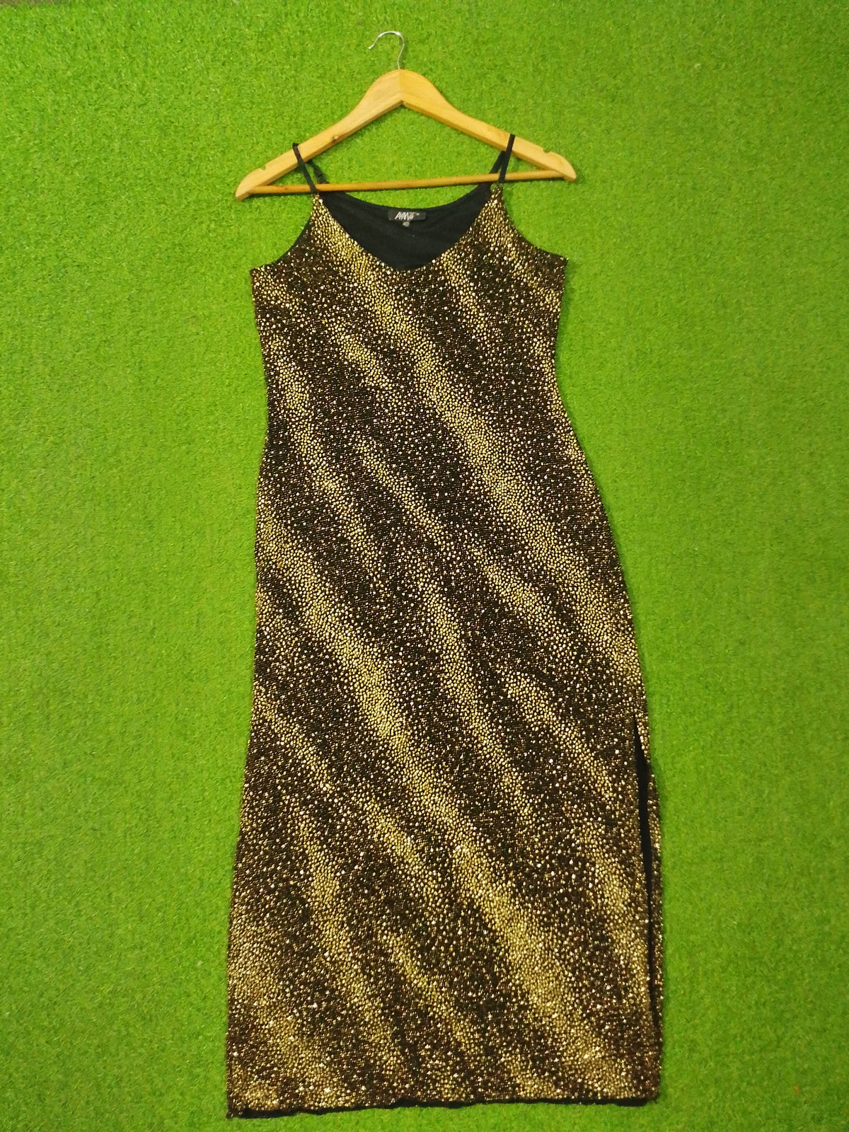 A sleeveless black slip dress with a gold glitter zebra-stripe pattern, hanging on a wooden hanger against a bright green textured background.