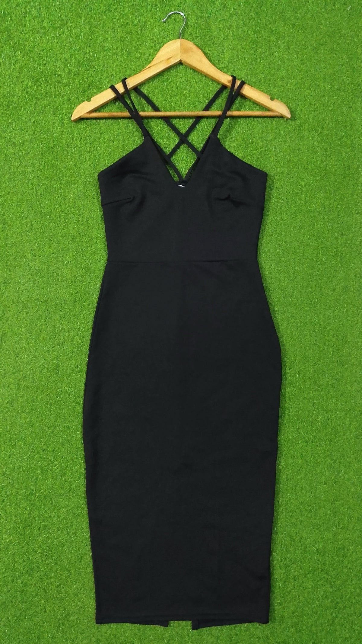 A black sleeveless bodycon dress with thin crisscross shoulder straps, hanging on a wooden hanger against a bright green textured background.