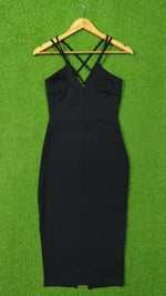 A black sleeveless bodycon dress with thin crisscross shoulder straps, hanging on a wooden hanger against a bright green textured background.
