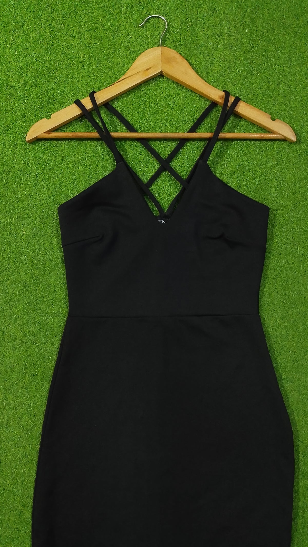 A black sleeveless dress with thin crisscross shoulder straps hanging on a wooden hanger against a bright green textured background.