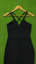 A black sleeveless dress with thin crisscross shoulder straps hanging on a wooden hanger against a bright green textured background.