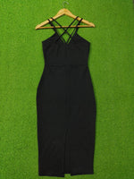 A black sleeveless bodycon dress with crisscross shoulder straps, hanging on a wooden hanger against a bright green artificial grass background.