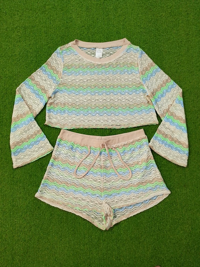 A two-piece girls' lounge set consisting of a long-sleeve cropped knit top and matching shorts, both featuring a beige and pink chevron pattern with pastel blue, green, and yellow stripes, laid flat on a green grass background.