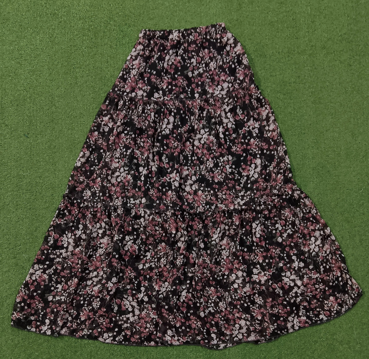 A black midi skirt with a dense pink and white floral print, laid flat on a green textured background.