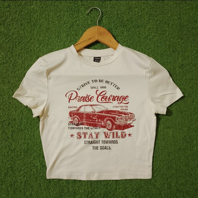 A white short-sleeve graphic T-shirt hanging on a wooden hanger against a green artificial turf background. The shirt features a red vintage car graphic and distressed red text reading 'Praise Courage', 'STAY WILD', and 'STRAIGHT TOWARDS THE GOALS'.