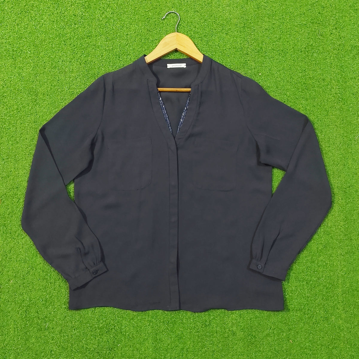 A dark charcoal gray long-sleeve button-up blouse with a split V-neckline, laid flat on a wooden hanger against a bright green artificial grass background.