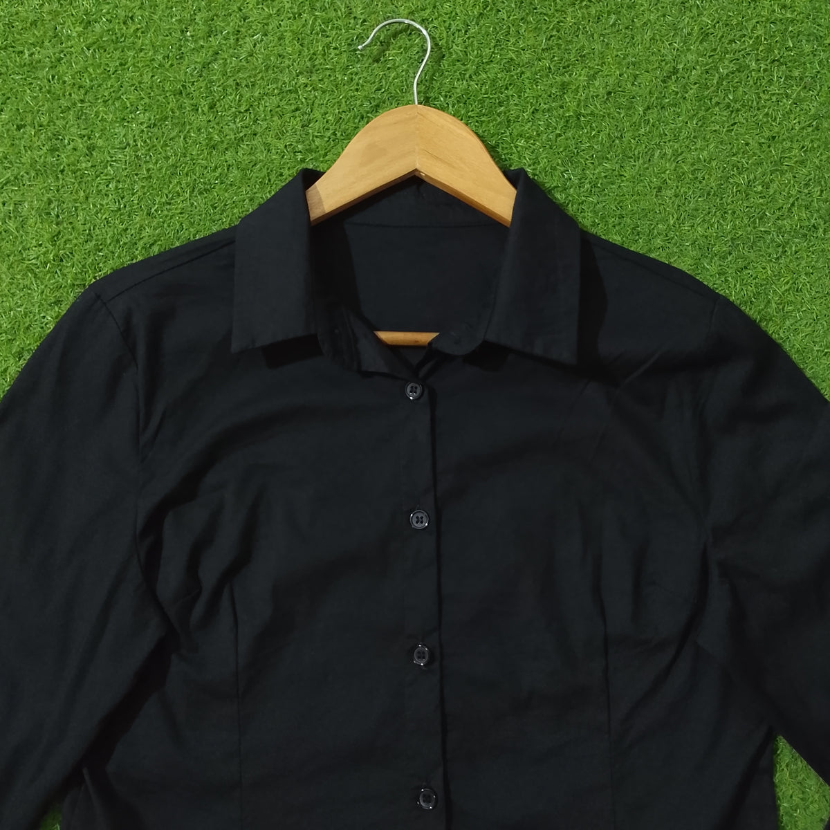 A black long-sleeve button-up shirt with a pointed collar, hanging on a wooden hanger against a bright green artificial grass background.