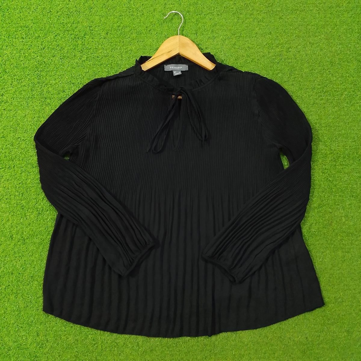 A black ribbed knit long-sleeve top with a drawstring neckline, laid flat on a bright green artificial turf background and hung on a wooden hanger.