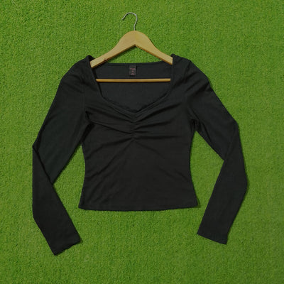 A black long-sleeve crop top with a ruched sweetheart neckline, hanging on a wooden hanger against a bright green grass-like background. - alternate view