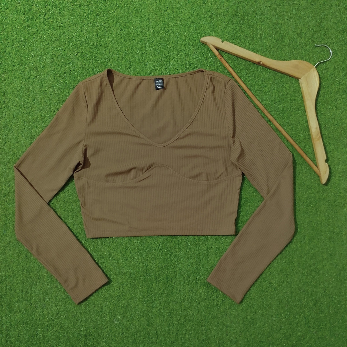A brown ribbed long-sleeve crop top laid flat on a green grass-like surface next to a wooden hanger.