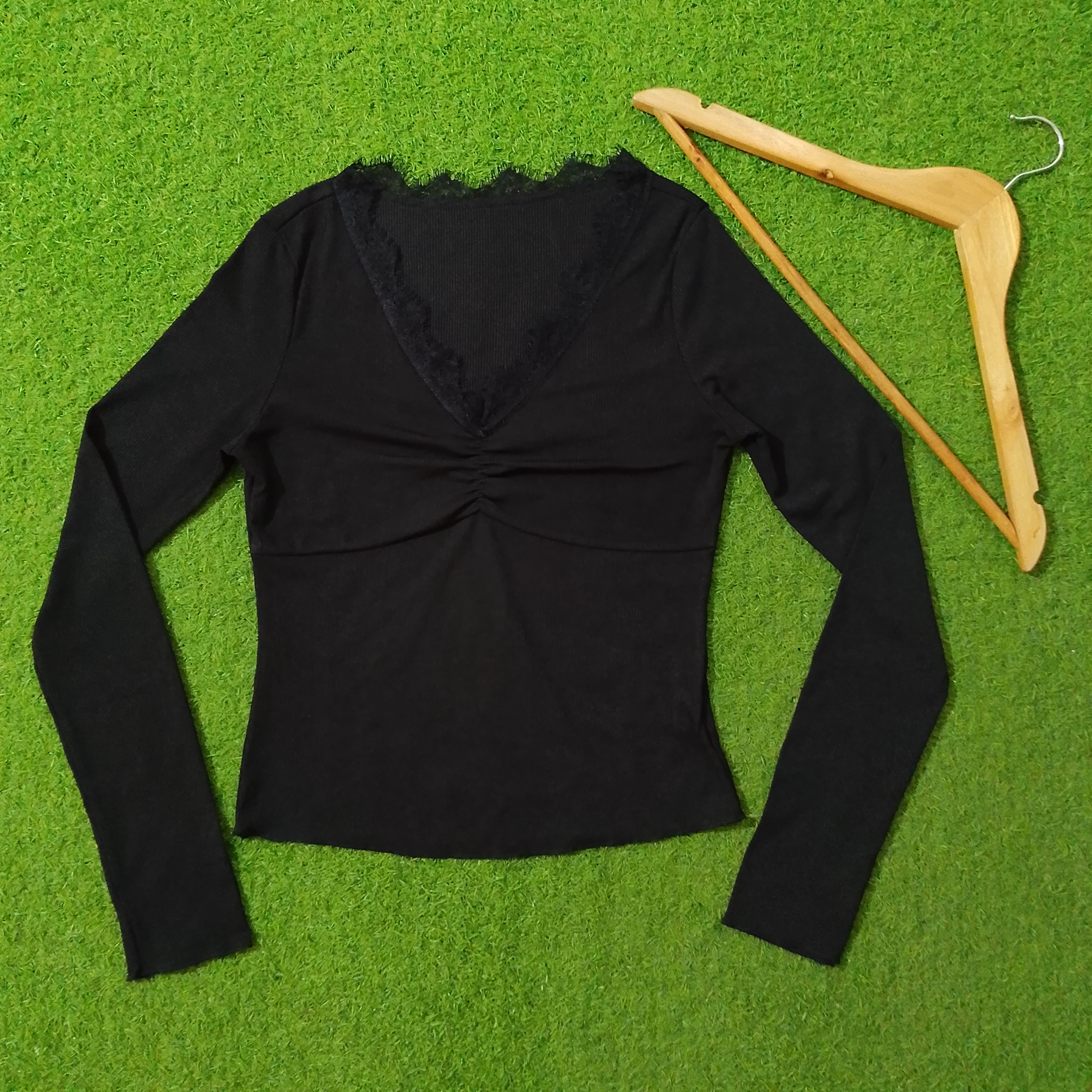A black long-sleeve V-neck top with a lace-trimmed neckline and gathered front, laid flat on a bright green textured surface next to a wooden hanger.