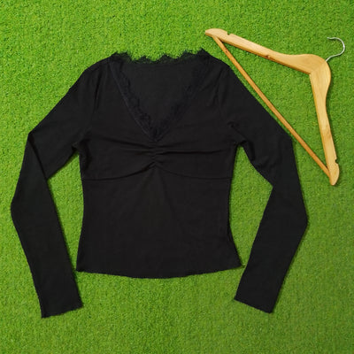 A black long-sleeve V-neck top with a lace-trimmed neckline and gathered front, laid flat on a bright green textured surface next to a wooden hanger.