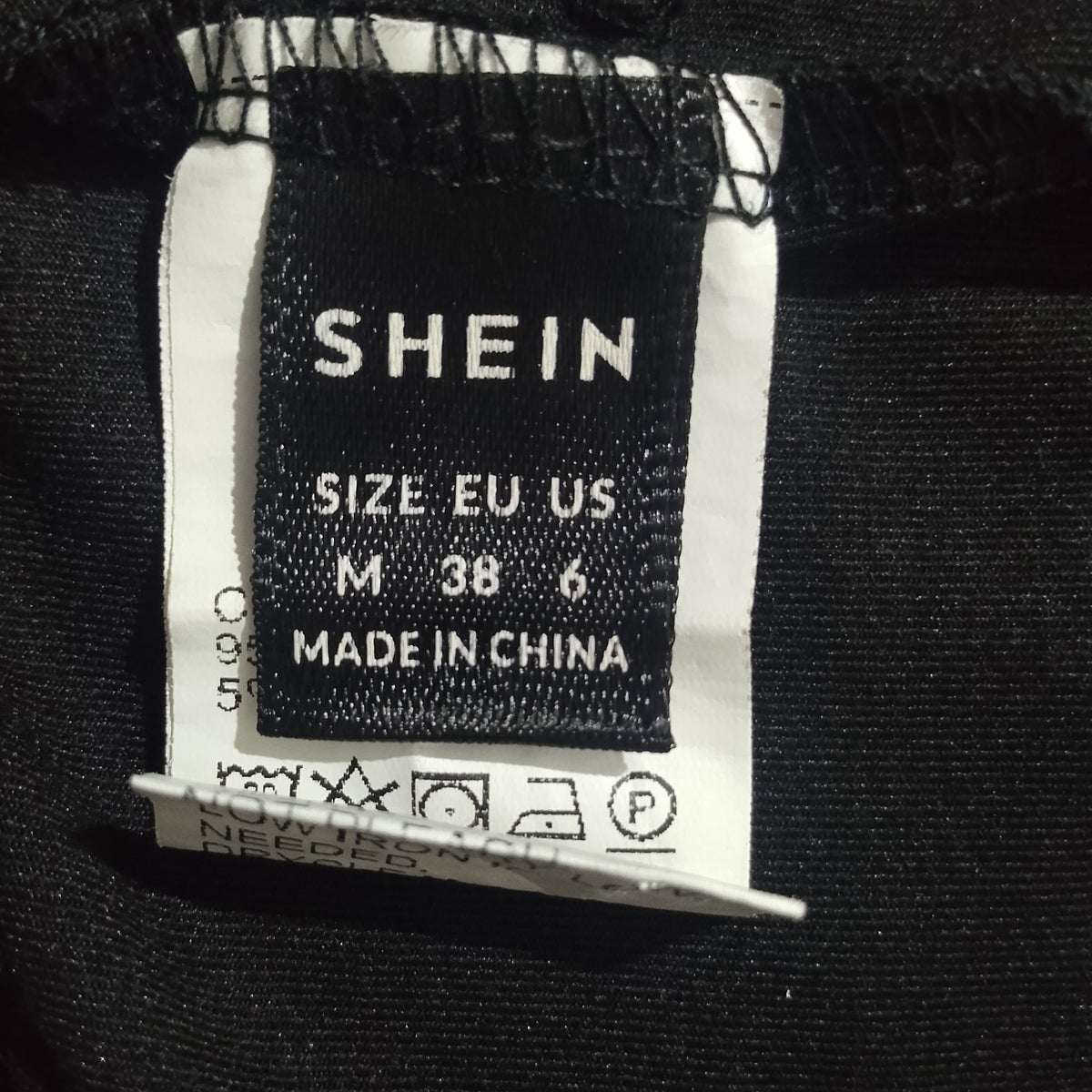 Close-up of a clothing size and brand label sewn onto black fabric. The black woven label reads SHEIN, size EU US M 38 6, MADE IN CHINA, alongside care instruction symbols on a white backing.
