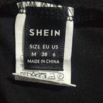Close-up of a clothing size and brand label sewn onto black fabric. The black woven label reads SHEIN, size EU US M 38 6, MADE IN CHINA, alongside care instruction symbols on a white backing.