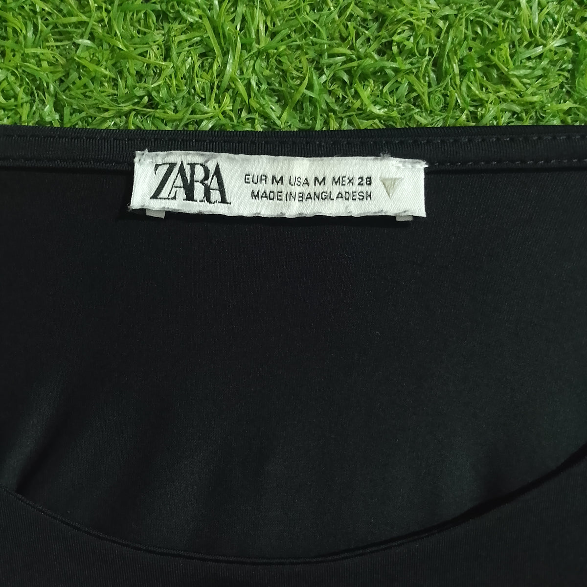 Close-up of a black garment with a white ZARA neck label, photographed against bright green grass.