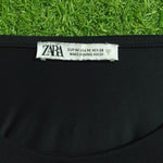 Close-up of a black garment with a white ZARA neck label, photographed against bright green grass.