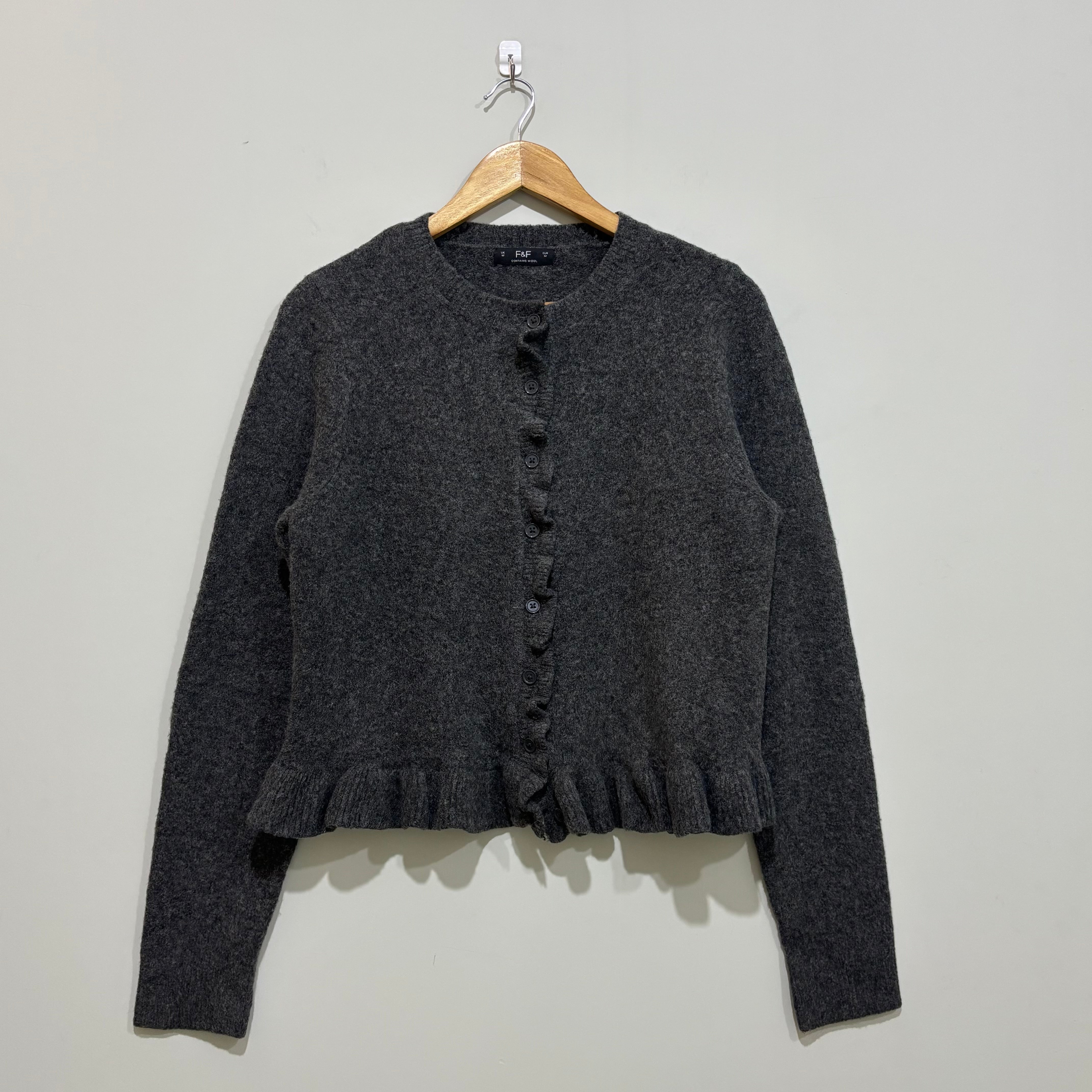 Charcoal grey long-sleeve cardigan with a round neckline, ruffled button-front trim and ruffle hem on a wooden hanger.