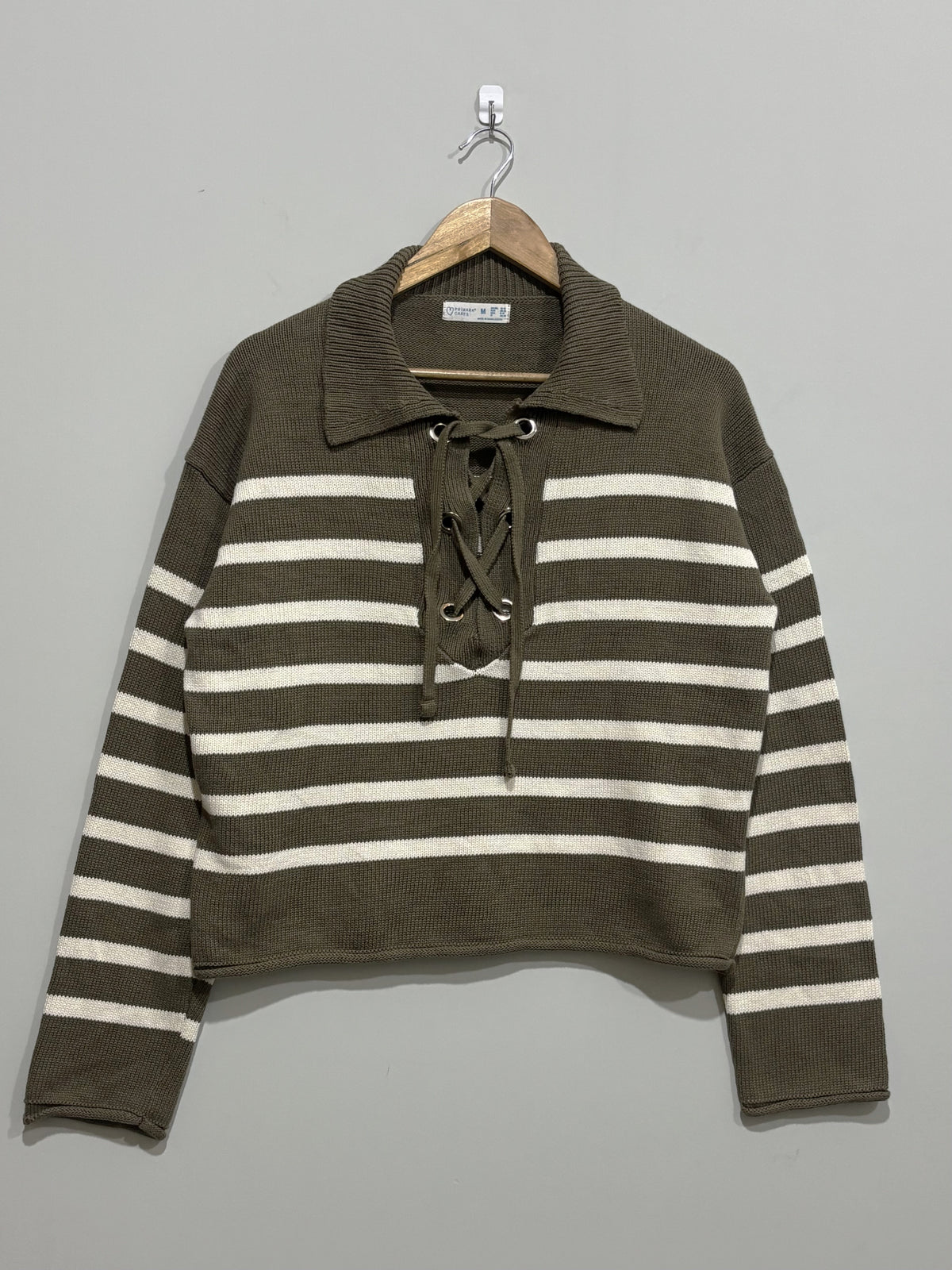 Olive green and cream horizontally striped pullover with a wide collar, lace-up neckline and long sleeves on a wooden hanger.