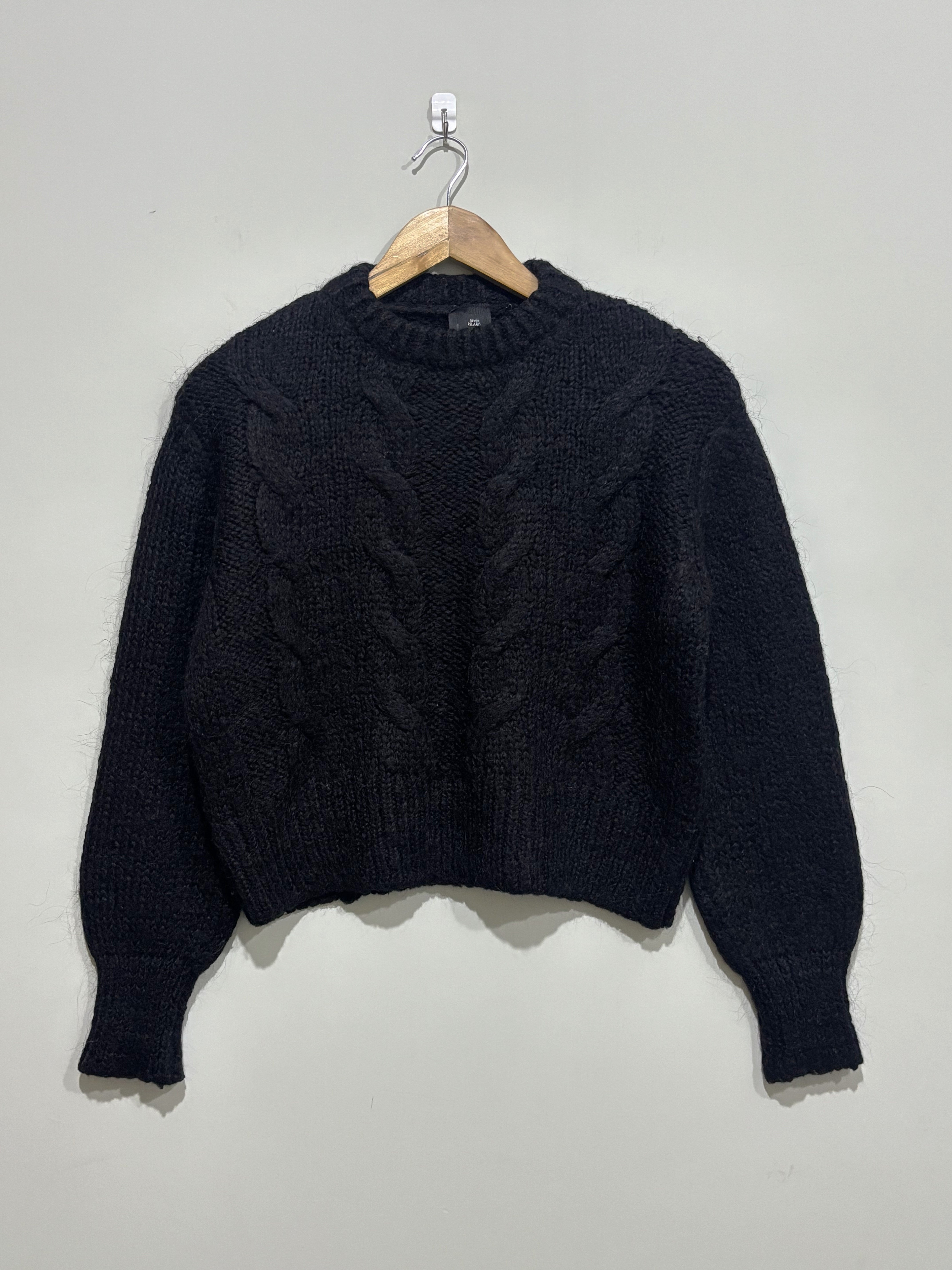 Black cable-knit pullover with a crew neckline, full balloon sleeves and ribbed cuffs and hem on a wooden hanger.