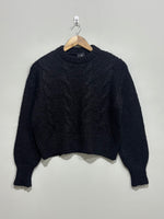 Black cable-knit pullover with a crew neckline, full balloon sleeves and ribbed cuffs and hem on a wooden hanger.