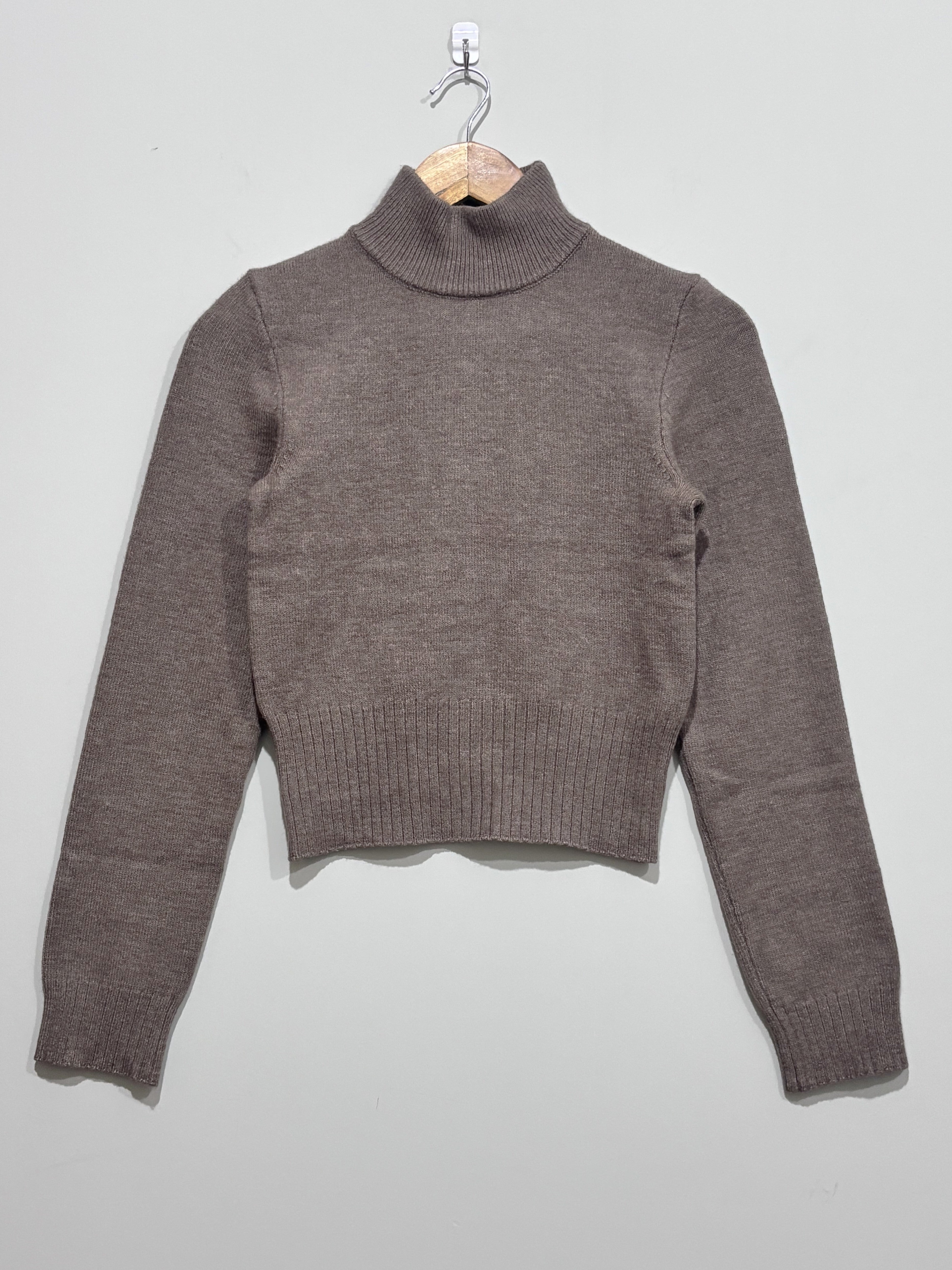 Taupe knitted pullover with a ribbed high neck, long sleeves and a deep ribbed hem, hanging on a wooden hanger.