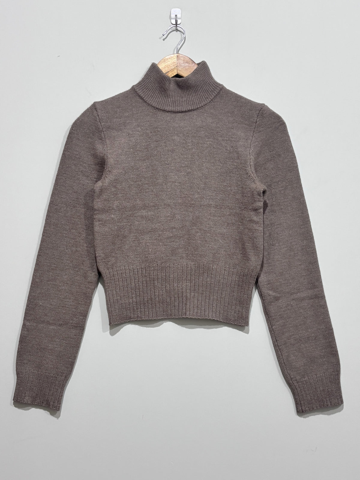 Taupe knitted pullover with a ribbed high neck, long sleeves and a deep ribbed hem, hanging on a wooden hanger.