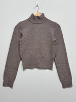 Taupe knitted pullover with a ribbed high neck, long sleeves and a deep ribbed hem, hanging on a wooden hanger.