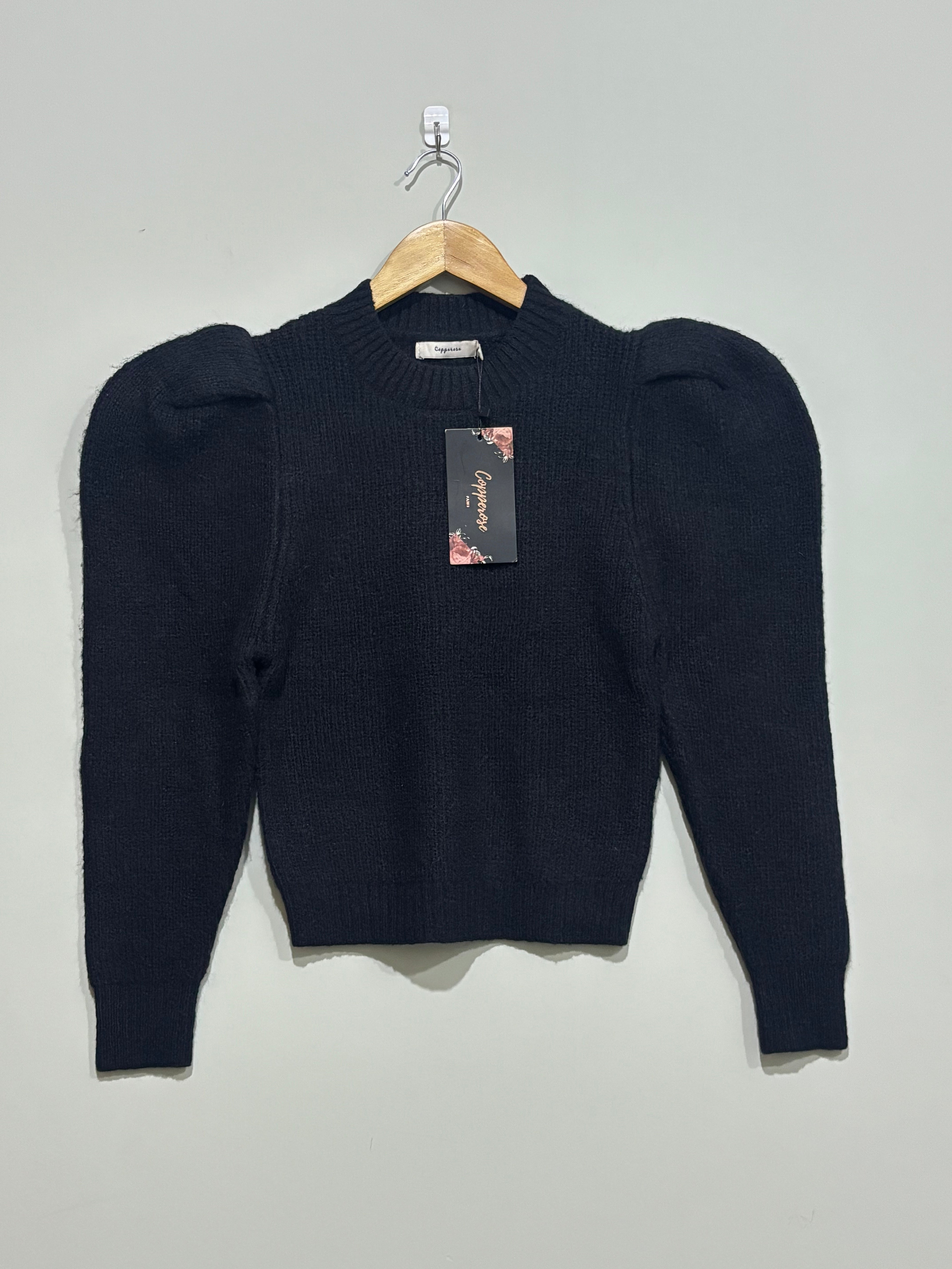 Black ribbed knit pullover with a round neckline, pronounced puff shoulders and long tapered sleeves, shown on a wooden hanger with a Glamorous hangtag.