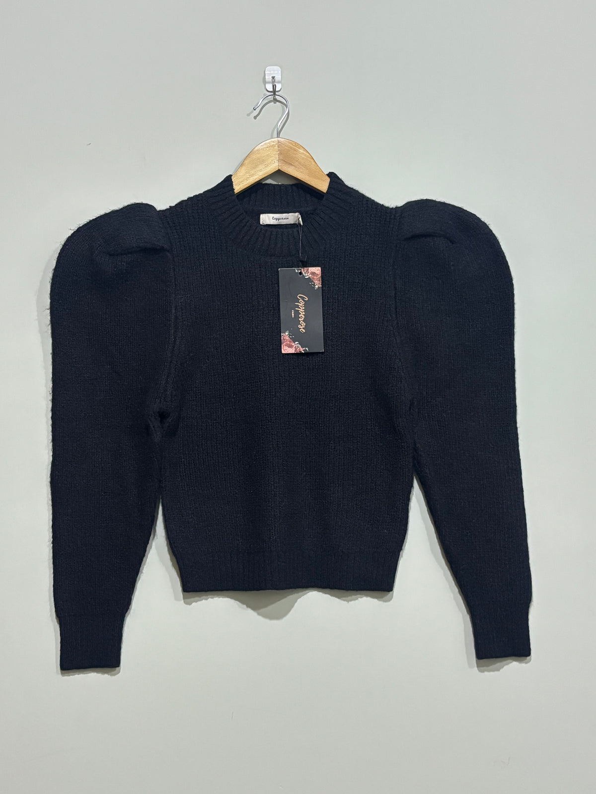 Black ribbed knit pullover with a round neckline, pronounced puff shoulders and long tapered sleeves, shown on a wooden hanger with a Glamorous hangtag.