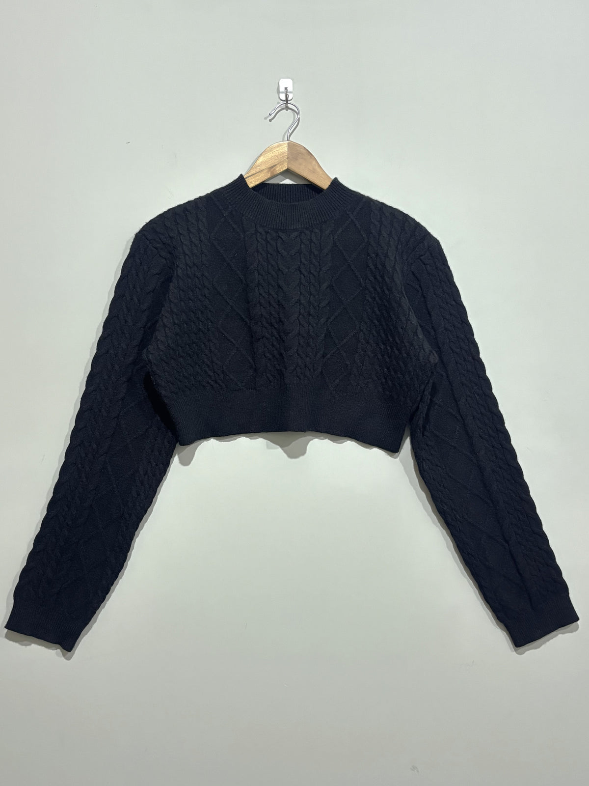 Black cropped cable-knit winter pullover with a round neckline, long sleeves and ribbed cuffs and hem, hanging on a wooden hanger.