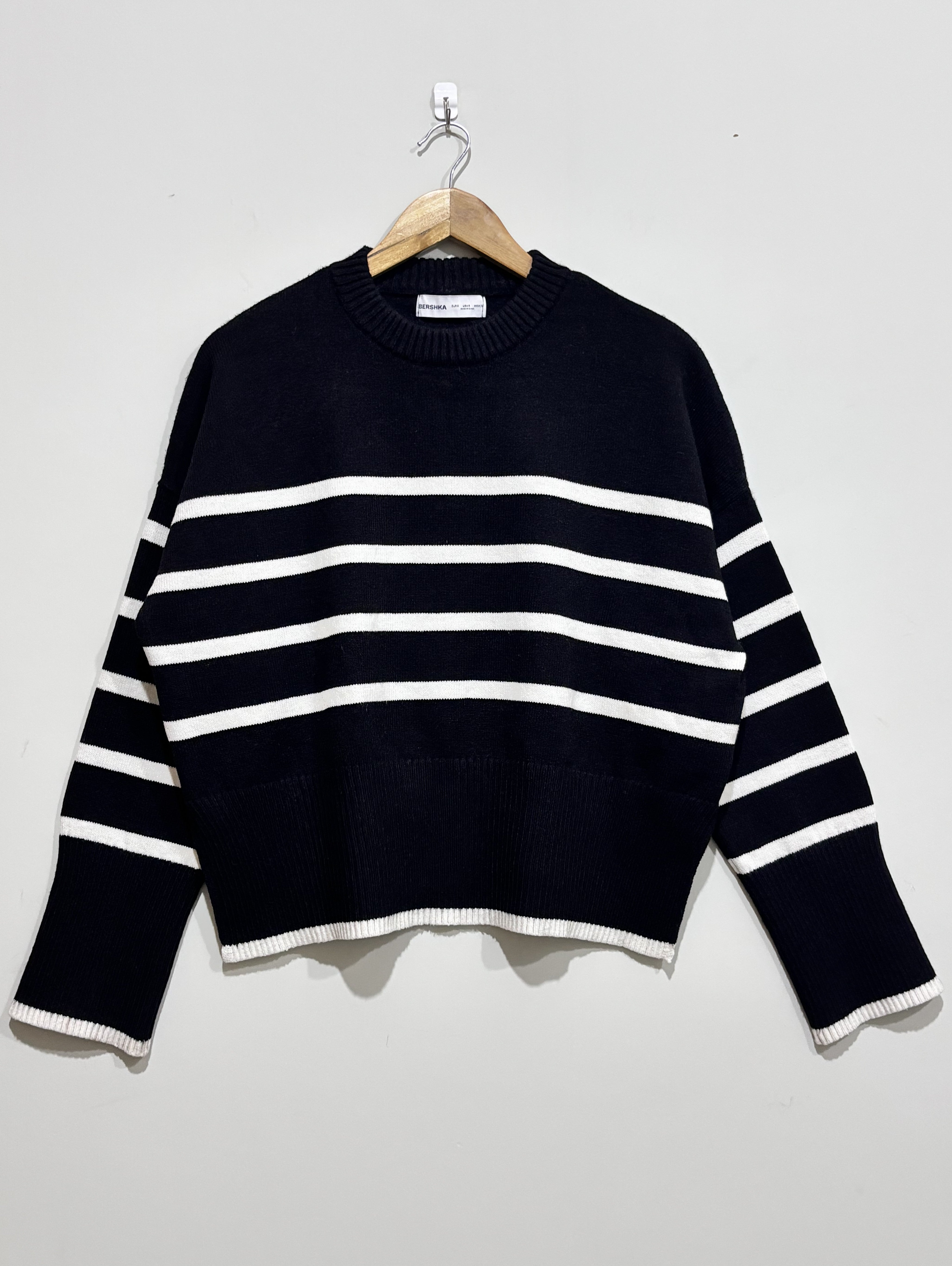 Black knit pullover with white horizontal stripes, a crew neckline, dropped shoulders and white edging at the cuffs and hem, on a wooden hanger.