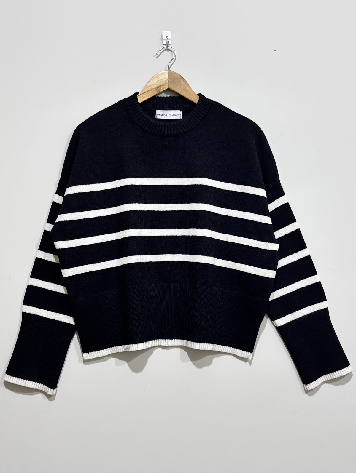 Black knit pullover with white horizontal stripes, a crew neckline, dropped shoulders and white edging at the cuffs and hem, on a wooden hanger.
