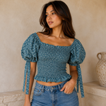 A woman wearing a smocked blue top with a white floral pattern, puff sleeves, and tie details, paired with blue jeans, standing against a textured beige wall.