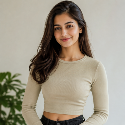 A woman with long dark brown hair wearing a beige ribbed long-sleeve crop top and black jeans, standing against a light beige wall with a green plant in the background.