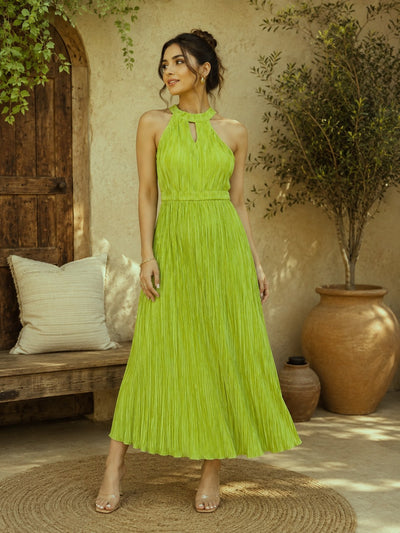 A woman modeling a bright lime green halter-neck midi dress with a pleated skirt, standing in a rustic outdoor patio setting with a wooden bench, pillows, and large terracotta pots.