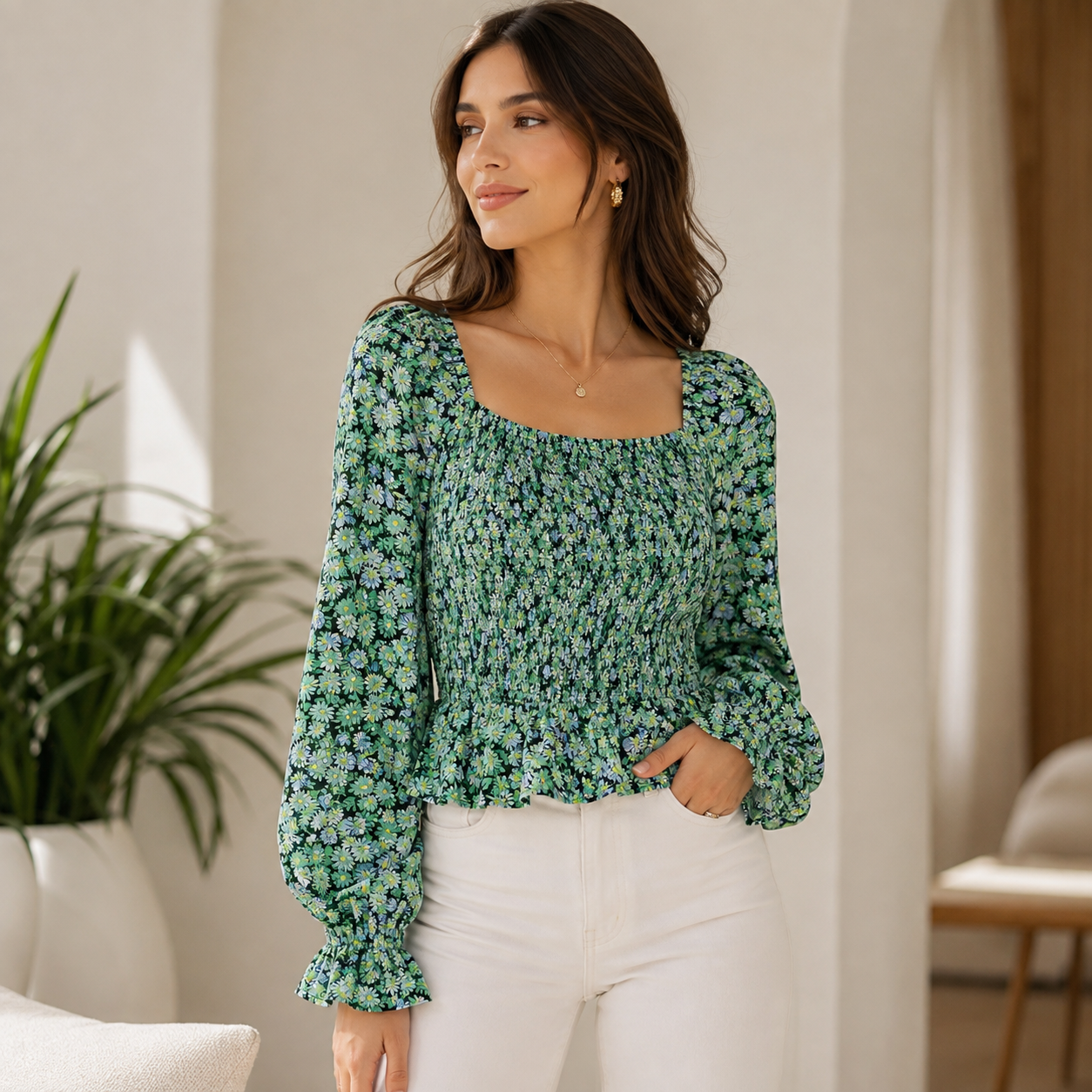 A woman wearing a green floral long-sleeve blouse with a smocked bodice and square neckline, paired with white pants, standing in a bright indoor setting.