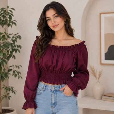 A woman wearing a burgundy off-the-shoulder long-sleeve crop top with a smocked waist and ruffled neckline, paired with light blue jeans, standing in a brightly lit indoor setting.