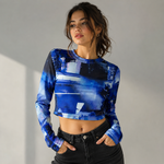 A woman modeling a blue abstract print long-sleeve crop top paired with black jeans, standing against a neutral gray wall with soft sunlight shadows.