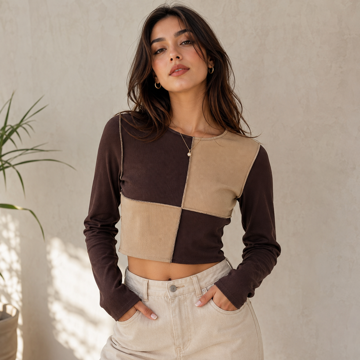 A woman wearing a long-sleeve color-block crop top in dark brown and beige corduroy, paired with light beige jeans, standing against a textured beige wall.