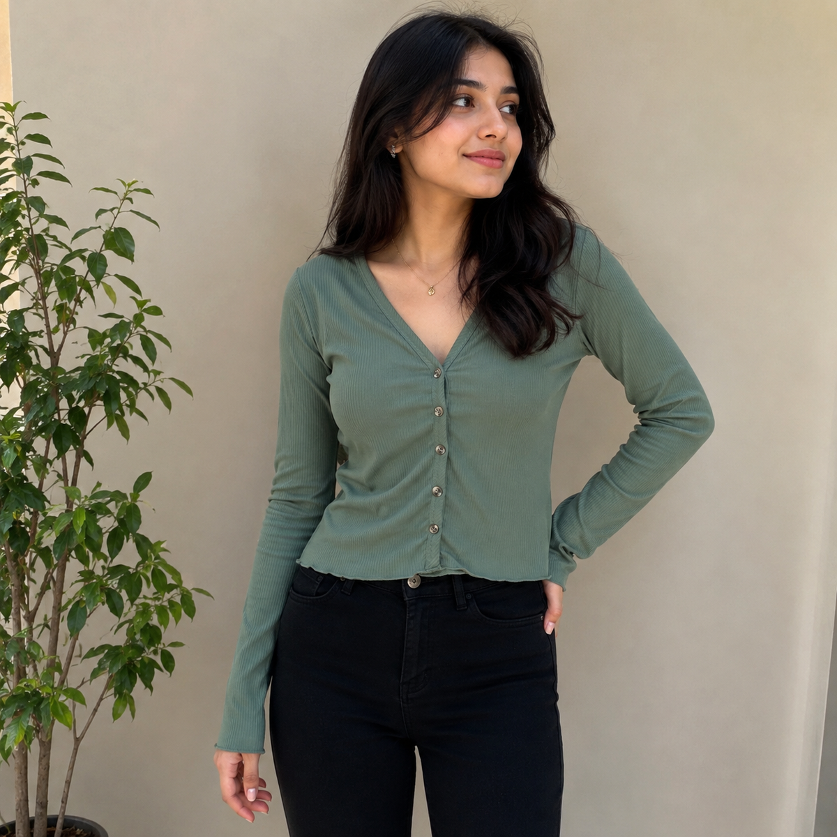 A woman wearing a sage green long-sleeve button-up top and black jeans, standing against a beige wall with a potted plant on the left.