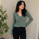 A woman wearing a sage green long-sleeve button-up top and black jeans, standing against a beige wall with a potted plant on the left.