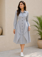 A woman wearing a light gray long-sleeve button-down shirt dress with a draped asymmetrical hem, standing in front of a beige stucco wall next to a potted plant.