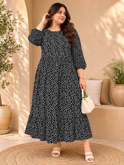 A woman models a black midi dress with a white floral pattern, featuring three-quarter sleeves and a tiered skirt, set against a warm beige outdoor patio backdrop with potted plants and a woven rug.