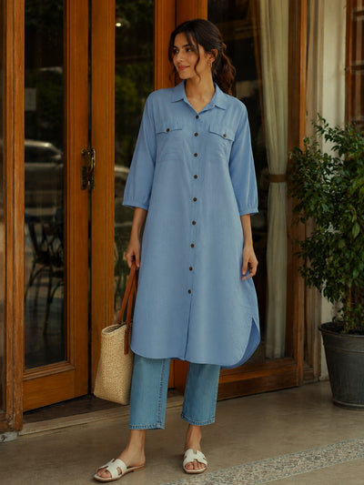 A woman wearing a light blue button-front tunic shirt with three-quarter sleeves and light blue cropped jeans, standing in front of a wooden door and holding a woven tote bag.