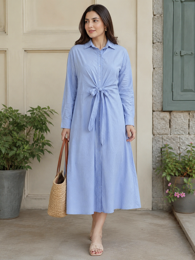 A woman models a light blue long-sleeve midi shirt dress with a tie waist, standing in front of a beige stucco wall with stone and green accents. She is holding a woven straw handbag.