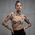 A woman modeling a long-sleeve mock-neck crop top with a brown and beige wavy swirl pattern, paired with black bottoms, set against a textured gray concrete wall.