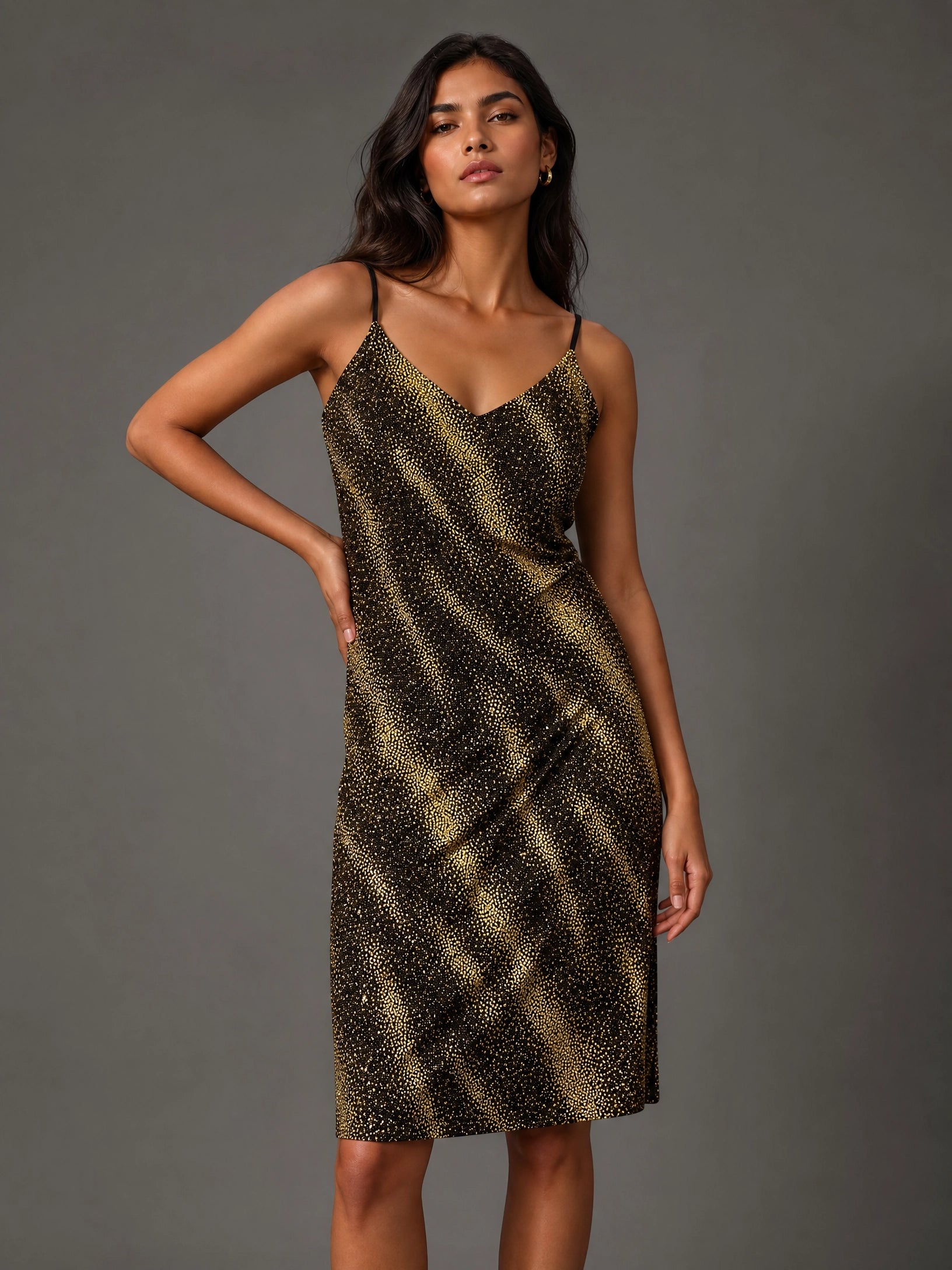 A woman modeling a sleeveless spaghetti-strap midi dress with a black and gold abstract spotted pattern, posed against a solid dark gray studio background.