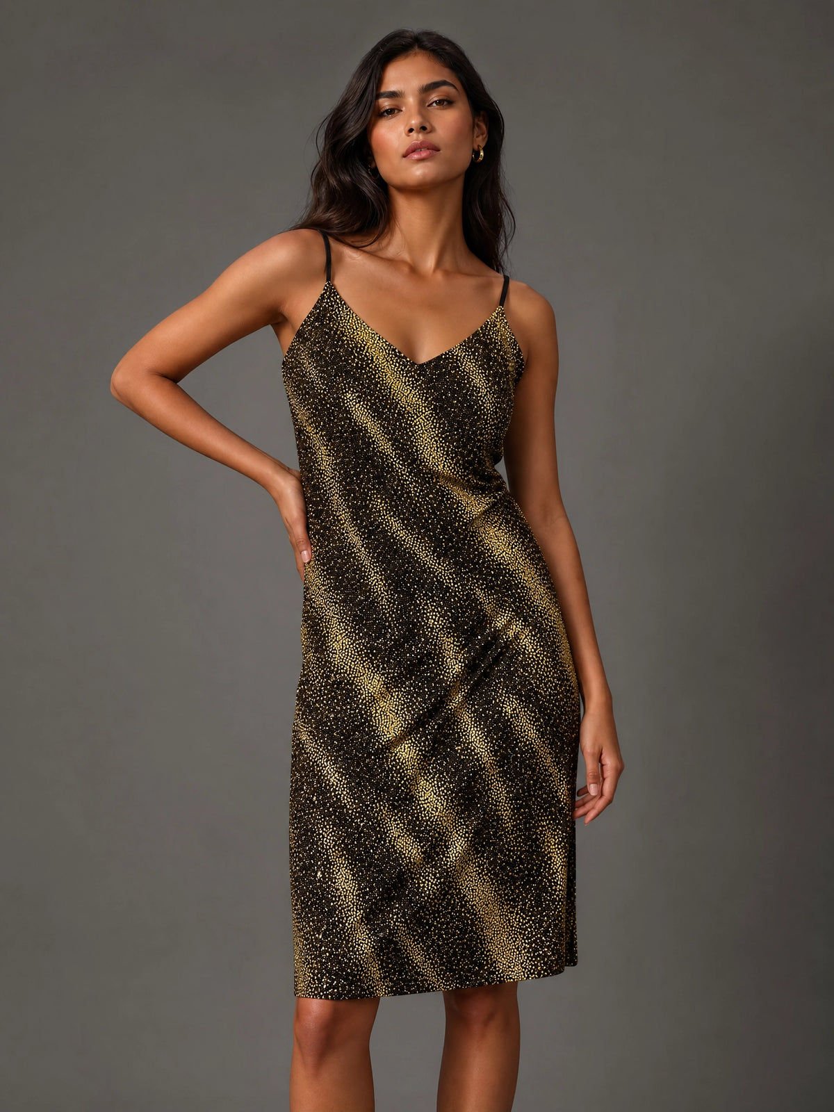 A woman modeling a sleeveless spaghetti-strap midi dress with a black and gold abstract spotted pattern, posed against a solid dark gray studio background.