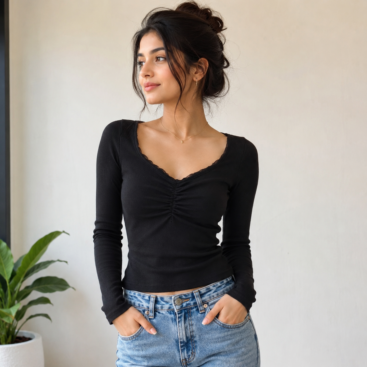 A woman wearing a black long-sleeve ruched top with a scalloped scoop neckline, paired with light blue jeans, standing against a neutral off-white wall.